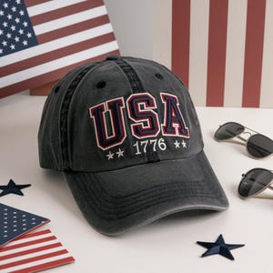 May include: A charcoal gray baseball cap with the word "USA" embroidered in red and navy blue, along with "1776" and two stars. The cap is a patriotic accessory.