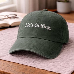 Könnte beinhalten: Dunkelgrüne Baseballkappe mit dem gestickten Text "He's Golfing." Die Kappe hat einen gebogenen Schirm und eine abgerundete Oberseite. Der Hut liegt auf einer hellvioletten Oberfläche.