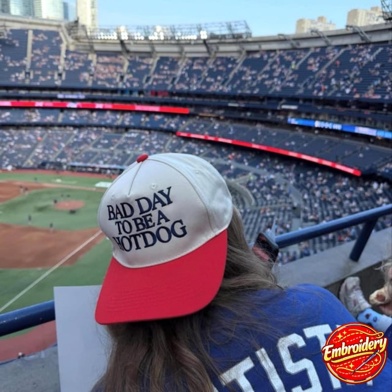 May include: A baseball cap with a white crown and a red brim. The front of the cap features the dark blue embroidered text "BAD DAY TO BE A HOTDOG". The cap is positioned in front of a large, blurred stadium with rows of seating and a baseball field.
