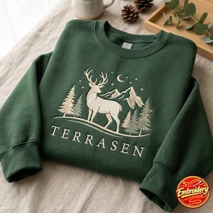 Puede incluir: Una sudadera de cuello redondo en color verde bosque oscuro con un diseño bordado de un ciervo en un paisaje montañoso con pinos, una luna creciente y estrellas. La palabra TERRASEN está bordada en color crema claro bajo el gráfico.
