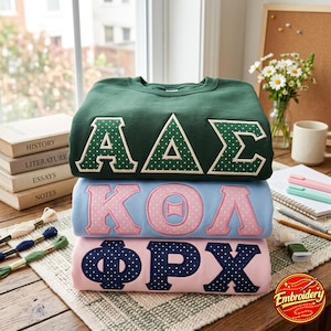 Könnte beinhalten: Ein Stapel aus drei Rundhals-Sweatshirts mit griechischen Buchstaben. Das oberste ist dunkelgrün mit weiß gepunkteten Buchstaben, das mittlere hellblau mit rosa gepunkteten Buchstaben und das unterste hellrosa mit dunkelblau gepunkteten Buchstaben.