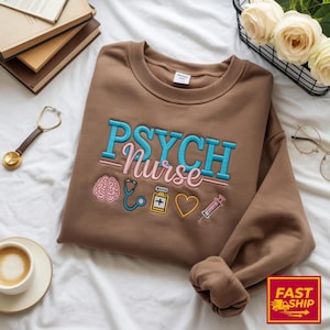 Op de afbeelding: Een bruine sweatshirt met de geborduurde tekst "PSYCH Nurse" in blauw en roze. Het ontwerp bevat een hersen, stethoscoop, medicijnfles, hart en spuit. De sweatshirt ligt op een witte ondergrond.