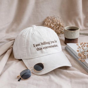 Op de afbeelding: Een witte baseball cap met de tekst "I am billing you for this conversation" in bruin. Een goudkleurige zonnebril met donkere glazen ligt ervoor. Een kopje koffie en een boek zijn op de achtergrond.
