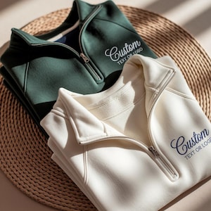 Könnte beinhalten: Zwei Sweatshirts mit Viertelreißverschluss, eines in Dunkelgrün und eines in Cremefarbe, sind ordentlich auf einer gewebten Matte gefaltet. Jedes Sweatshirt hat einen silbernen Reißverschluss und den gestickten Text "Custom TEXT OR LOGO" in Schreibschrift.