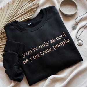 May include: Black sweatshirt with the embroidered text "You're only as cool as you treat people." The sweatshirt is displayed with a folded sleeve, a small bowl, and two necklaces.