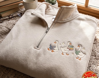 May include: A beige quarter-zip sweatshirt featuring an embroidered design of three white geese interacting with books. One goose sits reading, another carries a stack of books, and the third wears glasses while holding an open book, accented by small stars.