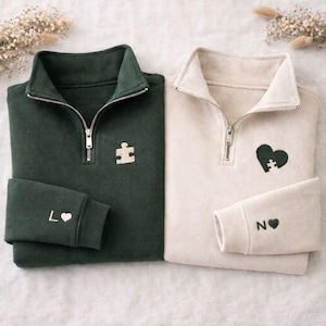 May include: Two quarter-zip sweatshirts, one dark green and one cream. Each has a puzzle piece design, one on the chest and one in a heart shape. The dark green sweatshirt has "L" and a heart on the cuff. The cream sweatshirt has "N" and a heart on the cuff.
