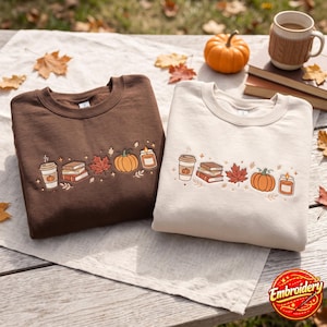 Può includere: Una felpa marrone scuro e una felpa color crema, ciascuna caratterizzata da una fila orizzontale ricamata composta da un bicchiere di caffè, una pila di libri, una foglia d'acero, una zucca e una candela. I disegni sono identici su entrambi.