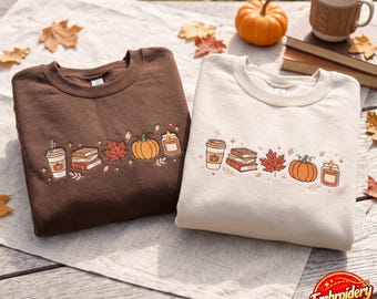May include: A dark brown sweatshirt and a cream sweatshirt, each featuring an embroidered horizontal row of a coffee cup, a stack of books, a maple leaf, a pumpkin, and a candle. The designs are identical in color and composition on both garments.