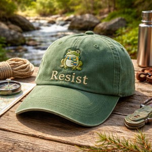 Könnte beinhalten: Grüne Baseballkappe mit einer gestickten Frosch-Applikation, die einen Schal trägt. Das Wort "Resist" ist darunter gestickt. Die Kappe liegt auf einer Holzoberfläche mit Kompass, Seil und Wasserflasche im Hintergrund.