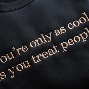 May include: Black sweatshirt with the embroidered text "you're only as cool as you treat people" in a light brown color. The sweatshirt has a crew neck and a soft, comfortable appearance. The text is centered on the front.