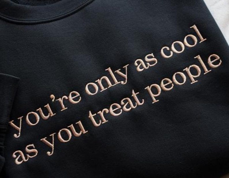 May include: Black sweatshirt with the embroidered text "you're only as cool as you treat people" in a light brown color. The sweatshirt has a crew neck and a soft, comfortable appearance. The text is centered on the front.