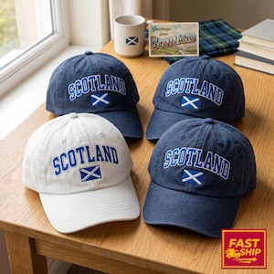Könnte beinhalten: Drei marineblaue Baseballkappen und eine weiße Baseballkappe, jeweils mit dem Wort SCOTLAND in blauer, geschwungener Schrift über einer kleinen gestickten schottischen Flagge. Die Kappen haben eine strukturierte Krone und einen gebogenen Schirm.