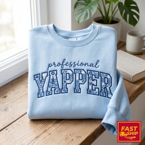 May include: A pale blue sweatshirt featuring the embroidered text "professional YAPPER" in blue. The word "YAPPER" is in a collegiate style font with floral detailing. The sweatshirt is folded on a wooden surface.