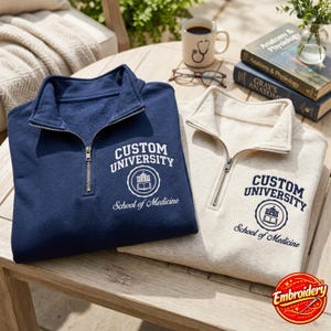 May include: A navy blue and a cream-colored quarter-zip sweatshirt, each featuring the white or dark blue text Custom University School of Medicine with a circular emblem of a building and an open book printed on the left chest area.