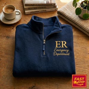 May include: Navy blue fleece pullover with a quarter-zip closure. The gold embroidered text reads "ER Emergency Department". The pullover is on a wooden surface, with a coffee cup and books in the background.