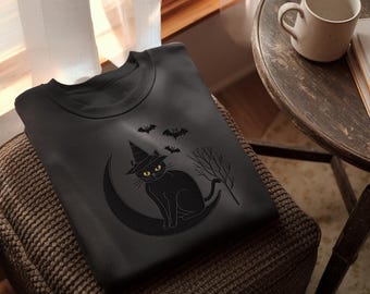 Puede incluir: Sudadera negra con un gato negro con sombrero de bruja sobre una luna creciente, con murciélagos y una escoba. La sudadera está sobre un otomán marrón cerca de una mesa de madera con una taza. El diseño es temático de Halloween.