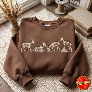 May include: A brown crewneck sweatshirt featuring an embroidered design of four goat skeletons in various poses, including walking, lying down, grazing, and standing on a rock. The embroidery is done in a light cream thread color.