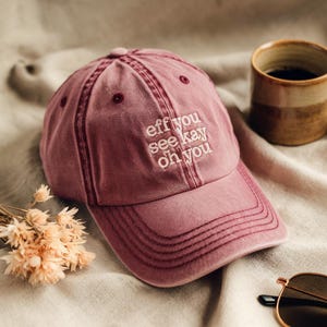 Könnte beinhalten: Verblasste rosa Baseballkappe mit dem gestickten Text "eff you see kay oh you" in Weiß. Die Kappe hat einen gebogenen Schirm und einen Used-Look mit sichtbaren Nahtdetails. Ein lässiges Accessoire.