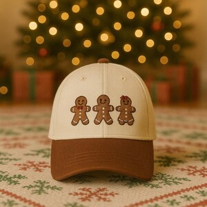 Pode incluir: Boné de beisebol com coroa creme e aba marrom. O boné apresenta um design bordado de três bonecos de gengibre. O fundo inclui uma árvore de Natal com luzes desfocadas e presentes embrulhados.
