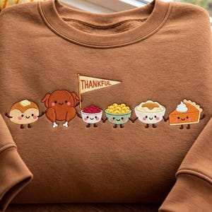 May include: A brown crewneck sweatshirt featuring an embroidered design of six anthropomorphic Thanksgiving food items holding hands. The characters include a pancake, a turkey, a bowl of cranberries, macaroni, mashed potatoes, and a slice of pumpkin pie. A pennant flag above the turkey reads THANKFUL.