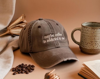 May include: A distressed brown baseball cap with the embroidered text "maybe coffee is addicted to me." The cap is next to a ceramic mug and coffee beans. The background is a neutral tan color.