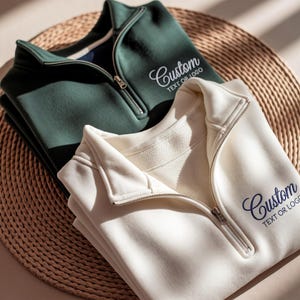 May include: A dark green and a cream-coloured quarter-zip sweatshirt, each featuring a silver metal zip and the embroidered text Custom TEXT OR LOGO on the upper left chest. The garments are folded and stacked, showcasing the collar and zip detail.