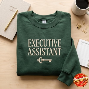 May include: A dark forest green crewneck sweatshirt featuring the embroidered text "EXECUTIVE ASSISTANT" in a light cream colour, positioned above a small, matching embroidered key icon centred on the chest of the garment.