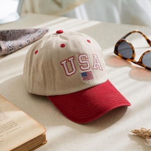 Könnte beinhalten: Eine beige Baseballkappe mit rotem Schirm und roten Ösen. Auf der Vorderseite befindet sich der gestickte Text USA in Rot mit weißer Umrandung, platziert über einer kleinen gestickten amerikanischen Flagge. Die Kappe hat eine feste Krone.