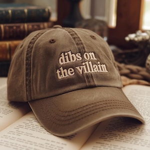 Może przedstawiać: Brązowa czapka bejsbolowa z białym haftem "dibs on the villain". Czapka leży na otwartej książce. Kapelusz ma zakrzywiony daszek i klasyczny sześciopanelowy design.