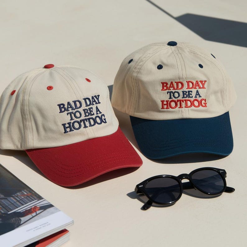 May include: Two baseball caps with cream-colored crowns and embroidered text reading BAD DAY TO BE A HOTDOG. One cap features a red brim and red eyelets, while the other has a dark blue brim and dark blue eyelets. Black sunglasses are included.