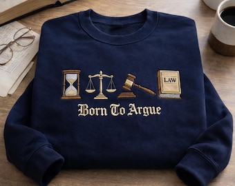 May include: A navy blue sweatshirt featuring embroidered law symbols: an hourglass, scales, a gavel, and a book with "LAW" on it. The words "Born To Argue" are embroidered below in a serif font.