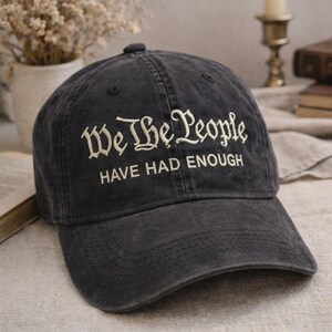 May include: A dark gray baseball cap with the embroidered text "We The People HAVE HAD ENOUGH" in white. The hat has a curved brim and a fabric strap closure. The cap is made of a worn, distressed material.