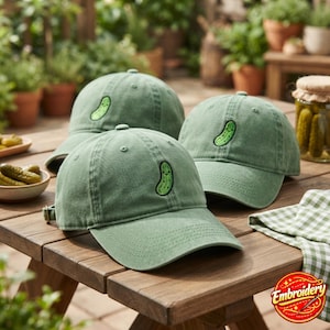 May include: Three sage green baseball caps made of cotton, each featuring a small, embroidered green pickle character with a smiling face on the front panel. The caps have a curved brim, a structured crown, and an adjustable metal buckle strap.