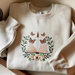Könnte beinhalten: Cremefarbenes Sweatshirt mit einem gestickten Design mit einem offenen Buch, einem Schwert, Schmetterlingen und floralen Elementen. Das Design ist in Braun-, Orange-, Grün- und Cremetönen gehalten. Das Sweatshirt hat einen Rundhalsausschnitt und lange Ärmel.