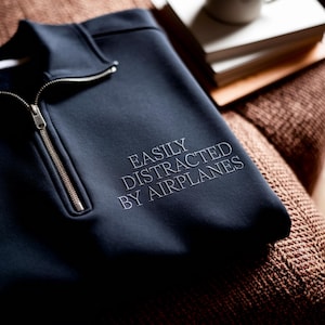 May include: Navy blue quarter-zip sweatshirt with the embroidered text "EASILY DISTRACTED BY AIRPLANES." The sweatshirt has a silver zipper and is resting on a brown textured surface.