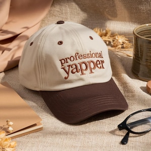 May include: A beige baseball cap with a brown brim and the embroidered text "professional yapper" in brown. The cap has a curved brim and a button on top. The hat is on a textured surface.