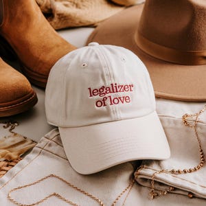 Può includere: Un cappellino da baseball color crema con la scritta ricamata "legalizer of love" in rosso. Il cappello è circondato da altri accessori moda, tra cui stivali in camoscio marrone, un fedora beige e collane d'oro.