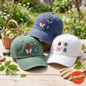 May include: Three baseball caps in blue, green, and white, each featuring embroidered designs of a red ladybug, a yellow and black bee, a blue beetle, and a pink butterfly. The caps are arranged on a wooden surface with a small woven basket.