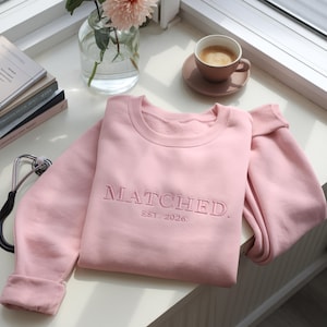 May include: A light pink sweatshirt with the word "MATCHED" embroidered on the front, along with "EST. 2026." The sweatshirt is folded on a white surface, next to a cup of coffee and a vase of flowers. The sweatshirt is a crew neck style.