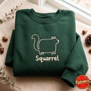 May include: A dark forest green crewneck sweatshirt featuring a light cream-colored embroidered outline of a squirrel and the text "Squarrel" centered on the chest. The garment has long sleeves with ribbed cuffs and a ribbed neckline.