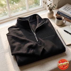 Op de afbeelding: Een zwart sweatshirt met een kwartritssluiting, een hoge kraag en een zilverkleurige metalen rits. Op de borst staat geborduurde tekst met de tekst BCBA Board Certified Behavior Analyst in een donkere, ton-sur-ton draad.