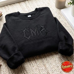 Op de afbeelding: Een zwart sweatshirt met ronde hals en zwarte borduursels op de borst. Het ontwerp bevat de letters CMA in een scherp, gotisch lettertype, met de tekst Certified Medical Assistant geborduurd in een kleiner, bijpassend zwart gotisch lettertype eronder.