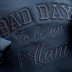 May include: Close-up of a navy blue sweatshirt with the embroidered text "BAD DAY To be an Alani" in a combination of block and cursive fonts. The sweatshirt's fabric has a soft, textured appearance.