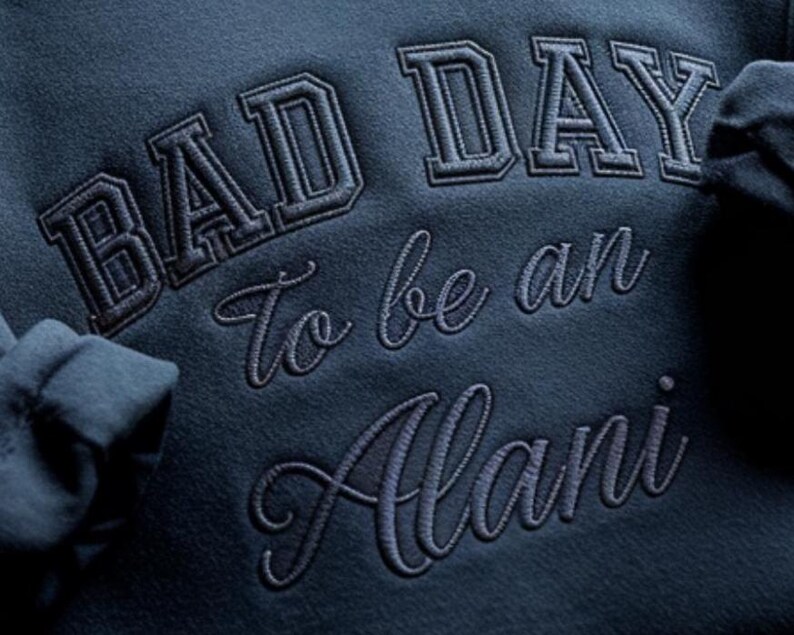 May include: Close-up of a navy blue sweatshirt with the embroidered text "BAD DAY To be an Alani" in a combination of block and cursive fonts. The sweatshirt's fabric has a soft, textured appearance.
