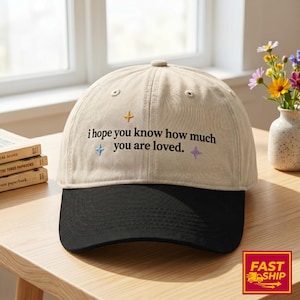 May include: A two-tone baseball cap with a beige crown and a black brim. The front of the crown features the embroidered black text "i hope you know how much you are loved." accented by three small, colorful star-shaped icons in yellow, blue, and purple.