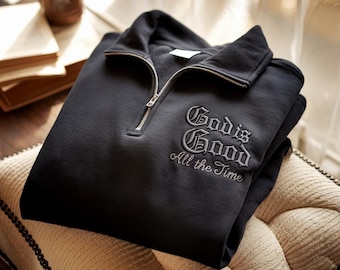Könnte beinhalten: Ein schwarzes Sweatshirt mit einem Viertel-Reißverschluss-Ausschnitt und dem gestickten Schriftzug "God is Good All the Time" in einer dekorativen Schriftart. Das Sweatshirt ist gefaltet und liegt auf einer gepolsterten Oberfläche.