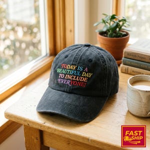 May include: A charcoal gray baseball cap with an embroidered message in multi-colored lettering that reads, "TODAY IS A BEAUTIFUL DAY TO INCLUDE EVERYONE!" The cap features a structured crown, a curved brim, and a button at the top center.