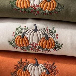 May include: Close-up of three folded sweatshirts in olive green, cream, and orange. Each sweatshirt features an embroidered design of a white pumpkin with orange pumpkins and greenery. The design is perfect for fall.