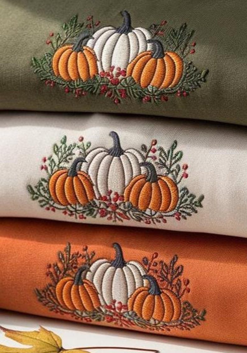 May include: Close-up of three folded sweatshirts in olive green, cream, and orange. Each sweatshirt features an embroidered design of a white pumpkin with orange pumpkins and greenery. The design is perfect for fall.
